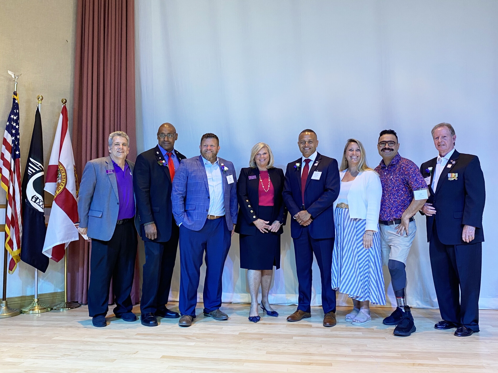 Group of eight adults in business attire posing on a stage with flags on the left, smiling for a formal photo.