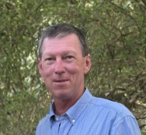 Middle-aged man in a blue striped shirt, outdoors with green foliage in the background.